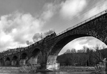 Historic steam train with old locomotive and nostalgic coaches passing the arch bridge viaduct in Witten Germany spanning over Ruhr river on a winter day. Vintage scenery black and white greyscale.