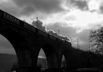 Historic steam train with old locomotive passing the arch bridge viaduct in Witten Germany spanning over Ruhr river. Vintage scenery and silhouettes back lit by low sun, black and white greyscale