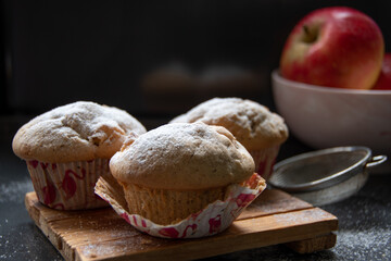 Rustikale Apfel Muffin frisch aus dem heimischen Offen . Das Zuckersieb liegt noch neben einen Holzbrett auf dem die Muffin angerichtet sind ,das ganze ist mit Zucker bestreut und auch noch köstliche 