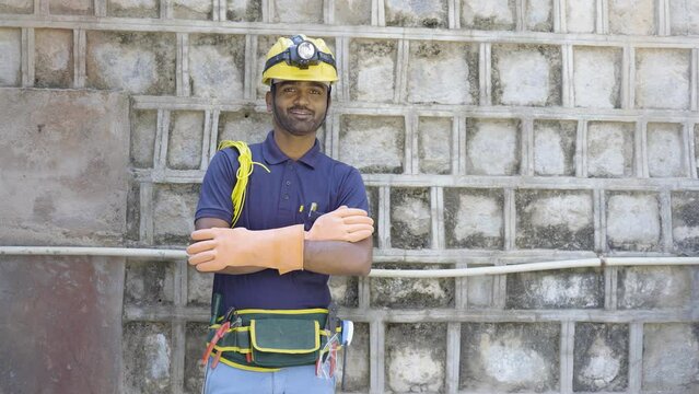 Electrician with tools and safety equipments looking at camera by crossing arms on industrial construction site - concpet of confident, blue collar jobs and maintenance service