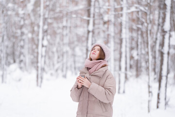 A young redheaded woman drinking tea from a thermos cup in a snow-covered winter forest.