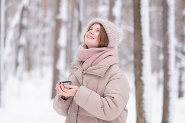 A young redheaded woman drinking tea from a thermos cup in a snow-covered winter forest.