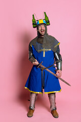 Full-length portrait of man, medieval warrior in armor, with sword wearing beer helmet and making...