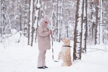 A young redheaded woman plays with her dog in the winter woods. A beautiful girl walks through a picturesque snow-covered park with a golden retriever. The concept of friendship with a pet.