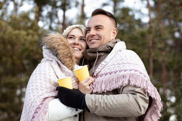two people in love. Hugging each other near a lot of spruce. Holding yellow cups with drink. Wear jeans, jacket.