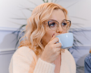 photo of young fascinating blond woman. Sitting on couch and drinking cup with tea. Look softly.