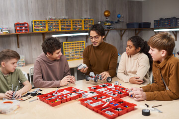 Portrait of smiling male teacher helping diverse group of teenage kids building robots in robotics...