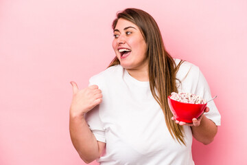 Young caucasian overweight woman holding a bowl of cereals isolated on pink background points with thumb finger away, laughing and carefree.