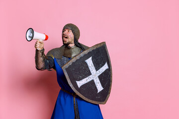 Portrait of man, medieval warrior or knight in armor with shield shouting in megaphone isolated...