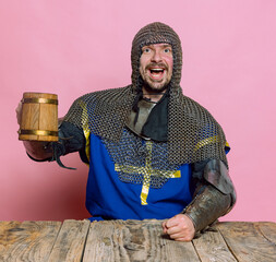 Portrait of excited man, medieval warrior, knight in special protection rising wooden beer mug...