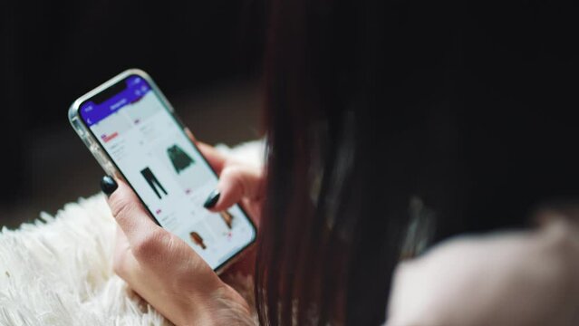 Online shopping concept. Young woman sitting on the bed and makes online shopping on a smartphone. Woman at Home Lying on Couch in Living Room Using Smartphone Buys in Internet Shop.