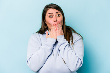 Young caucasian overweight woman isolated on blue background shocked covering mouth with hands.