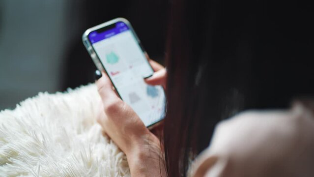 Online shopping concept. Young woman sitting on the bed and makes online shopping on a smartphone. Woman at Home Lying on Couch in Living Room Using Smartphone Buys in Internet Shop.