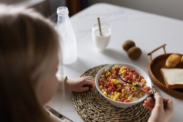 Cute little caucasian girl sitting at table on kitchen early morning and preparing breakfast with colorful cornflakes and milk. Kid enjoying life with healthful food, healthy lifestyle concept