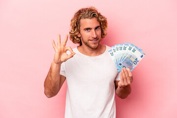Young caucasian man holding banknotes isolated on pink background cheerful and confident showing ok gesture.