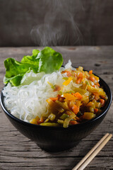 A plate of rice noodles stands on an old wooden table top. Rice noodles are most common in the cuisines of East, Southeast Asia and South Asia. The side dish consists of fried onions, carrots, paprika