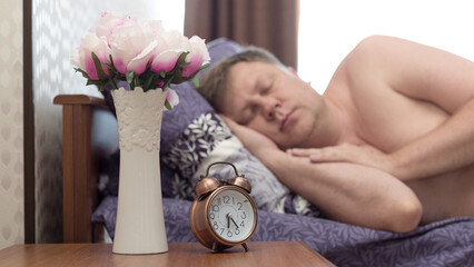 An adult man 40-45 years old is fast asleep in his bed without hearing the alarm clock