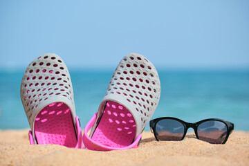 Obraz premium Closeup of clogs shoes and black protective sunglasses on sandy beach at tropical seaside on warm sunny day. Summer vacation concept