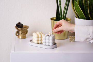 Female hand lighting a grey and off-white bubble candles on a concrete tray on white sideboard,...