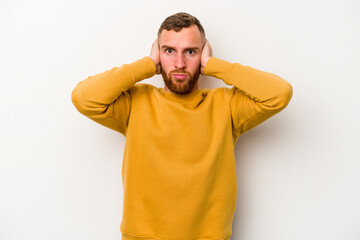 Young caucasian man isolated on white background covering ears with hands trying not to hear too loud sound.