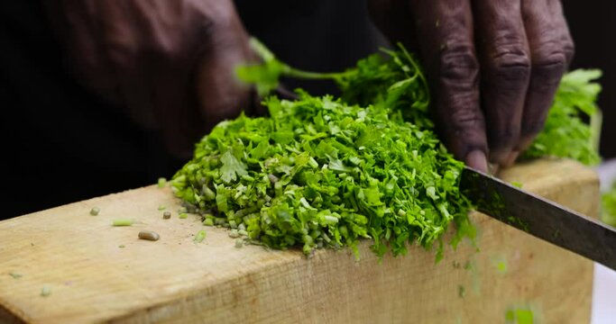 Close up shot of a chef finely chopping fresh organic coriander on a wooden board with a knife to prepare a meal. Natural green herb corriander, parsley, cilantro, mint, tulsi cutting for cooking. 