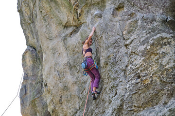 Young woman climbing steep wall of rocky mountain. Girl climber overcomes challenging route. Engaging in extreme sport concept