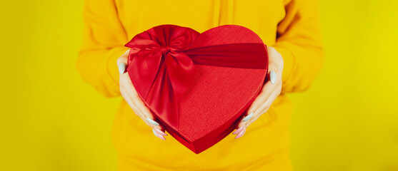 Body part of woman with red gift box in form of heart on yellow background. Unrecognizable person holding romantic gift with ribbon. Concept of present on valentine day and international womens day.