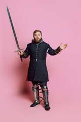 Full-length portrait of young bearded man, medieval warrior or archer in protection with sword isolated over pink background