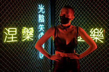 Young cyberpunk woman in black leather costume standing against bars with illuminated chinese...