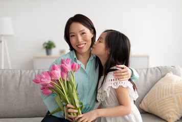 Cute Asian girl kissing cheerful grandmother, giving her flowers for Women's Day, greeting with birthday at home