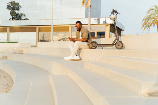 Photo with copy space of a black man using a mobile phone sitting on a stairs with a parked electric scooter.