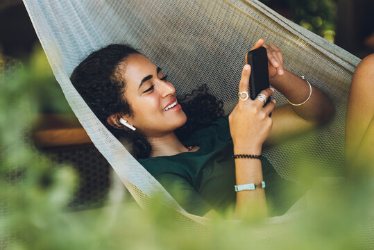 Beautiful brunette woman with laughing hair resting in a hammock with her cell phone and wireless headphones.