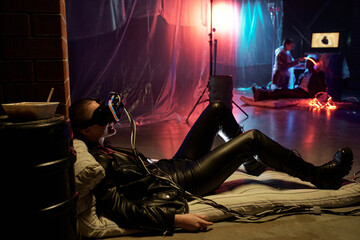 Young woman in leather costume lying on mattress in virtual reality glasses, people practicing artificial intellect in dark room in the background