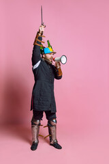 Fototapeta premium Full-length portrait of young man, medieval warrior or archer in beer helmet rising sword and shouting in megaphone isolated over pink background