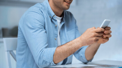 cropped view of smiling man in blue shirt messaging on cellphone in kitchen