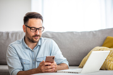 A serious young man working from home using a smartphone and notebook computer, male hands using a smartphone inside, a man at his workplace using technology. New normality concept