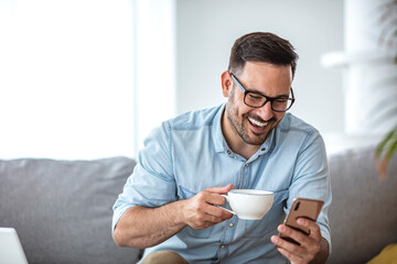 A smiling man makes a video call with friends while sitting on a sofa in his modern home. He uses a...