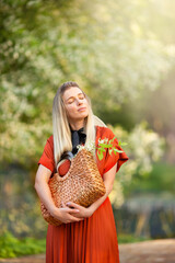 Young beautiful woman in a red dress enjoys a blooming spring garden. She holds a wicker bag with...