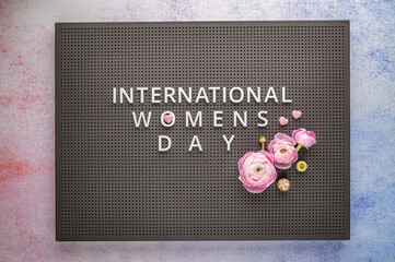 International womens day written on grey pegboard, ranunculus decorates the lettering on pastel background. Women's Day concept. Soft focus. Top view.