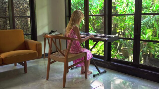 Blonde girl playing the piano in a beautiful modern home. Child is learning to play the electronic piano against the background of a glass wall and green garden.