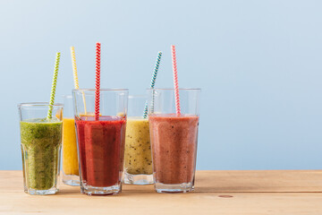 Set of multi-colored fruit smoothies in glasses with paper tubes on wooden table and blue background. Mango, kiwi, strawberry, apple summer drinks