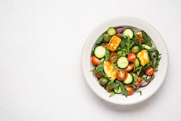 Halloumi salad with arugula, tomato, cucumber, onion and olives in bowl on light background. Mediterranean dish with fried cheese and vegetables