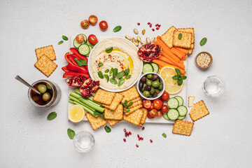 Hummus plate with vegetable snacks. Carrot, cucumber, tomato, celery sticks, dry crackers, olives and hummus bowl on marble tray and light background, top view