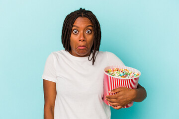 Young African American woman eating pop corns isolated on blue background  shrugs shoulders and open eyes confused.