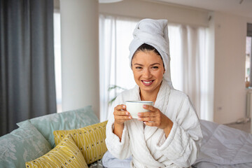 A lazy woman in a bathrobe lies on the bed and enjoys a sunny morning with a cup of aromatic coffee while relaxing in the comfortable bedroom of the hotel room