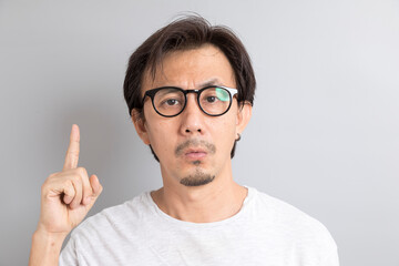 Adult asian man with glasses pointing fingers upward with curious face on grey background.