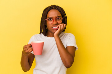 Young african american woman holding a mug isolated on yellow background  biting fingernails, nervous and very anxious.
