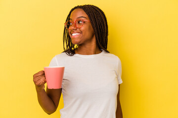 Young african american woman holding a mug isolated on yellow background  looks aside smiling, cheerful and pleasant.