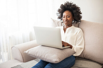 A beautiful young African girl with black curly hair in a sofa at home - checking online stores or searching for something on her laptop. He enjoys his free time.