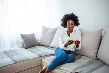 A beautiful African woman on a beige couch at home and shopping online, so comfortable and modern white clothes, denim pants, such a beautiful light interior design. The concept of online shopping